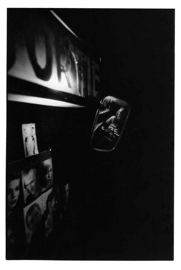 Photographie argentique noir et blanc de Simon Bezzi Batani montrant, dans un environnement sombre, le portrait par reflet de miroir d'une personne. Le miroir est entouré de portraits papier de grandes stars de cinéma américaines.